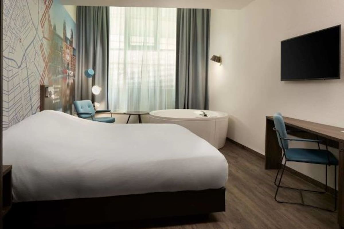 A hotel room with a large bed, a desk, a chair, a round table, and a TV on the wall.
