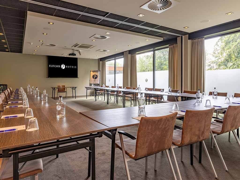 Un salón de conferencias con mesas y sillas largas, una pantalla de proyector y un amplio ventanal con cortinas.