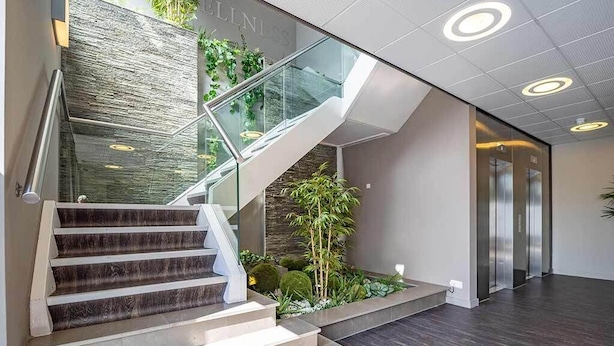 Un interior moderno con una escalera, barandas de vidrio y una zona con plantas.
