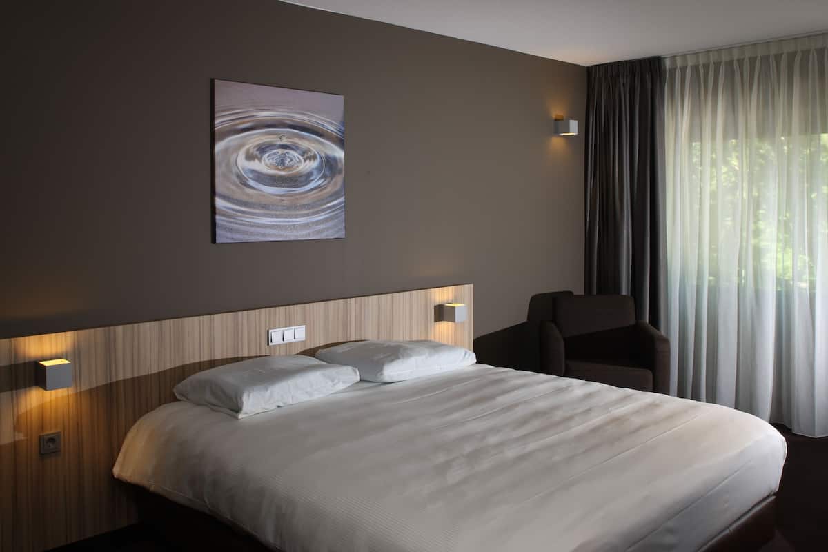 Ein Hotelzimmer mit einem großen Bett, einem Kopfteil mit integrierter Beleuchtung, einem Wandbild und einem Fenster mit Vorhängen.