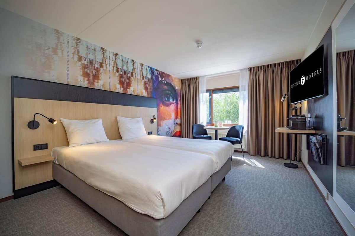 Ein Hotelzimmer mit einem großen Bett, einem Schreibtisch, einem Sessel, einem Fernseher und einem Fenster mit Vorhängen.