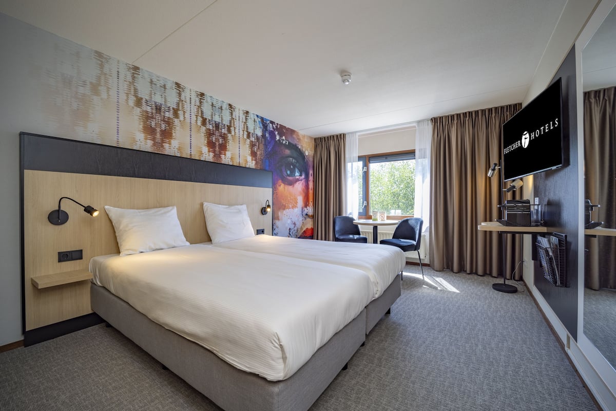 Ein Hotelzimmer mit einem großen Bett, einem Schreibtisch mit Stuhl, einem Fernseher und einem Fenster mit Vorhängen.