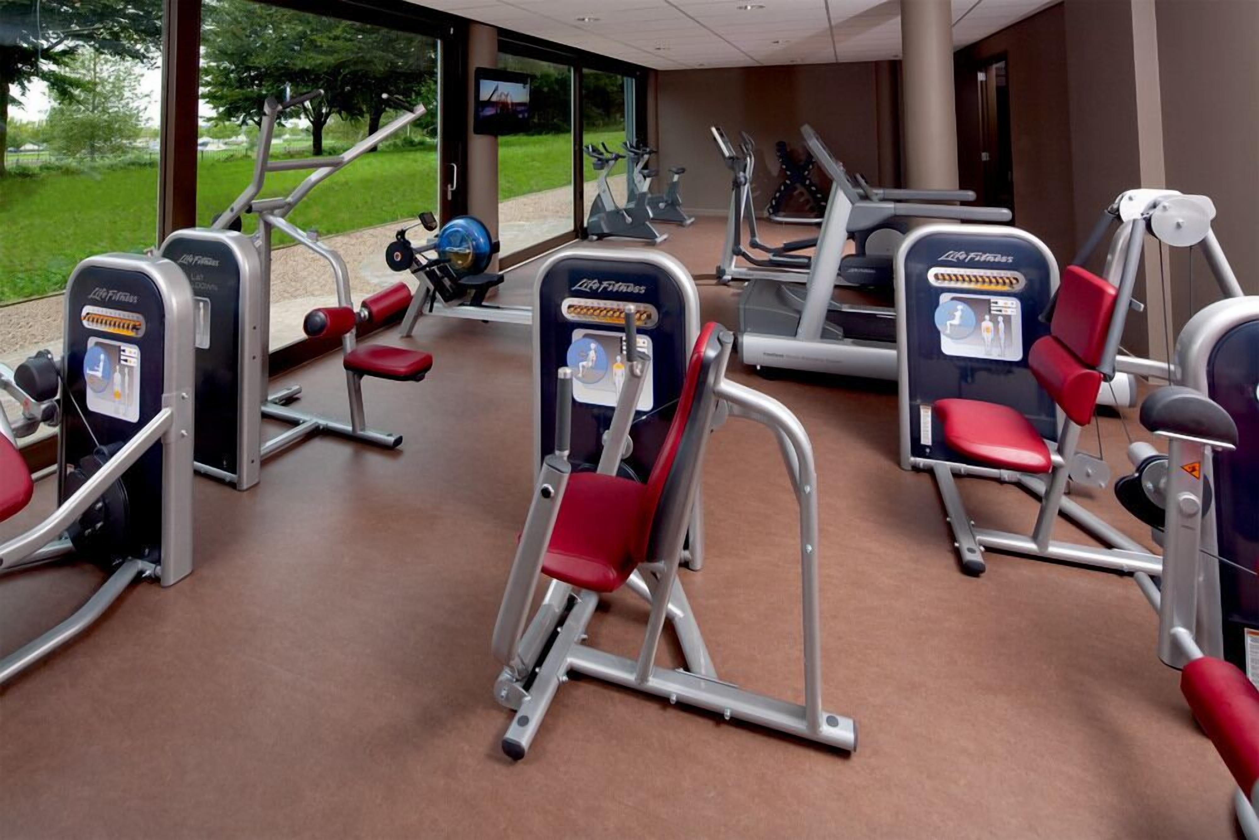 Ein Fitnessstudio mit verschiedenen Trainingsgeräten, darunter Crosstrainer und Kraftgeräte, in einem großzügigen Raum mit großen Fenstern und Blick ins Freie.