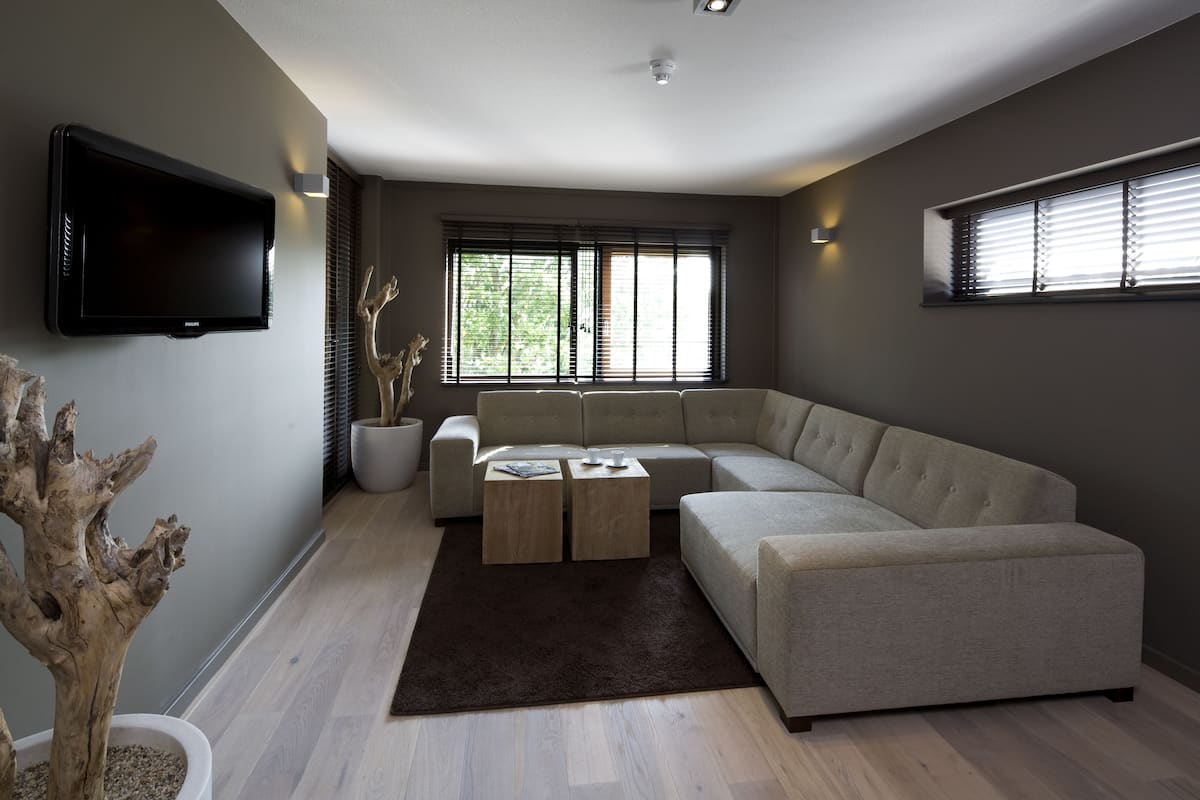 Ein modernes Wohnzimmer mit einer Eckcouch, einem flachbildfernseher an der Wand und einem großen Fenster mit Jalousien.