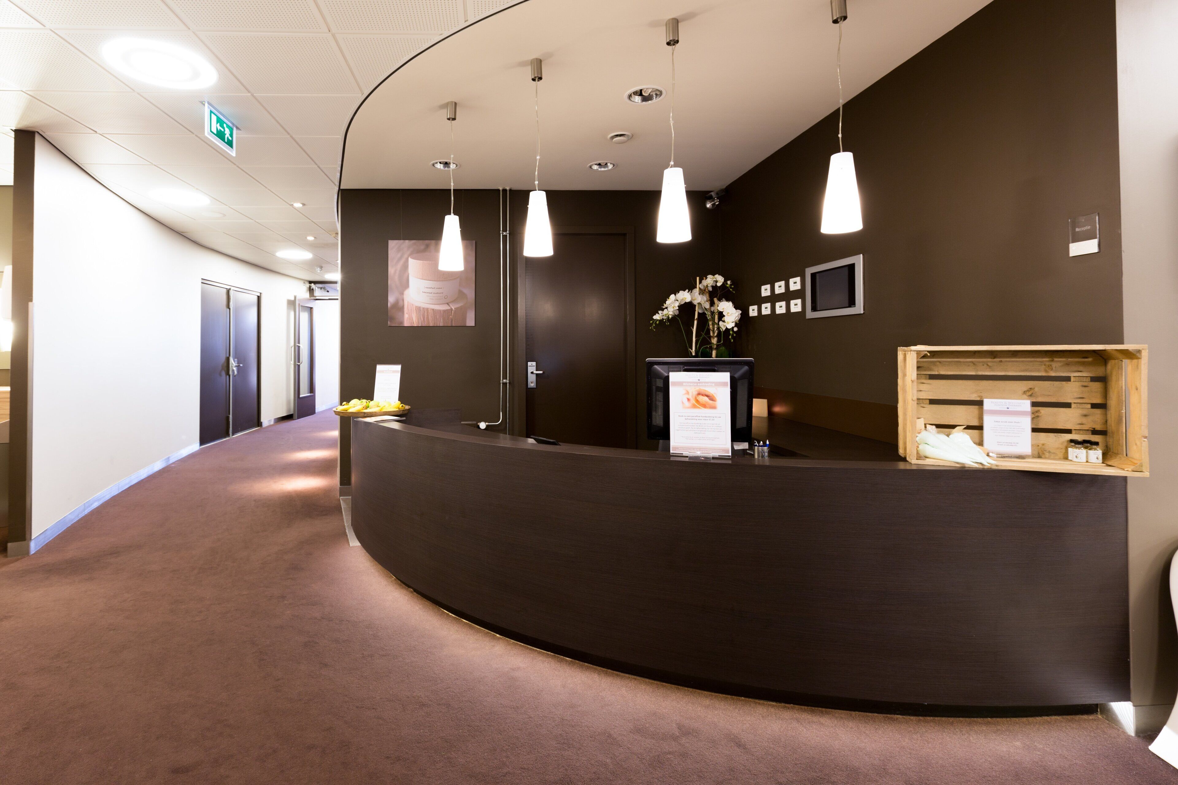 Een receptiegedeelte met een gebogen receptiebalie, hangende verlichting, een wandgemonteerde televisie en een houten displaykast met objecten.