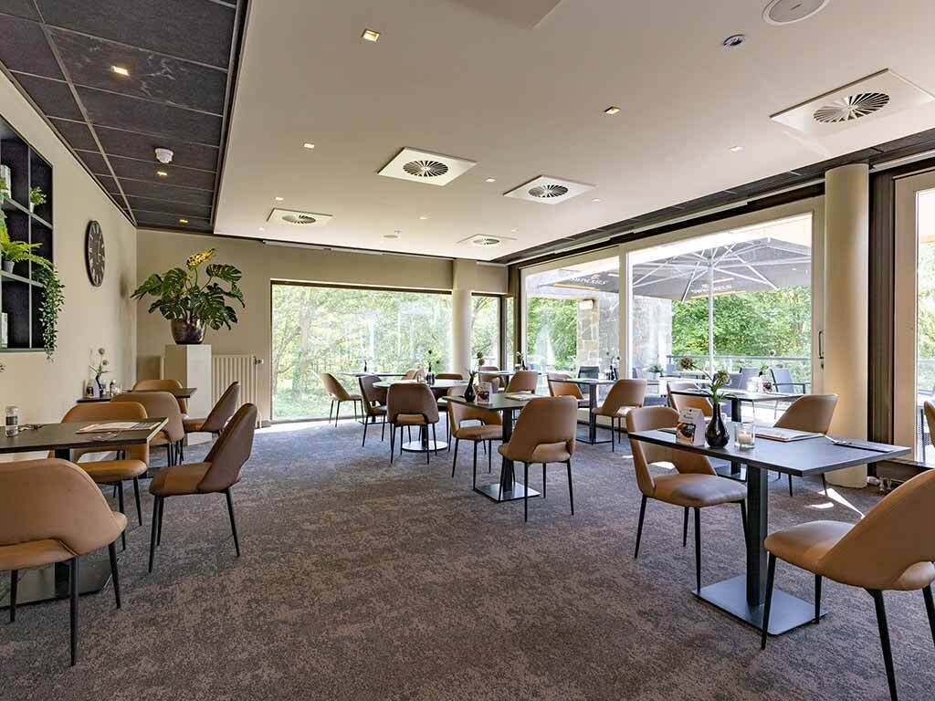 A spacious indoor dining area with multiple tables and chairs, large windows allowing natural light, and a ceiling with recessed lighting.