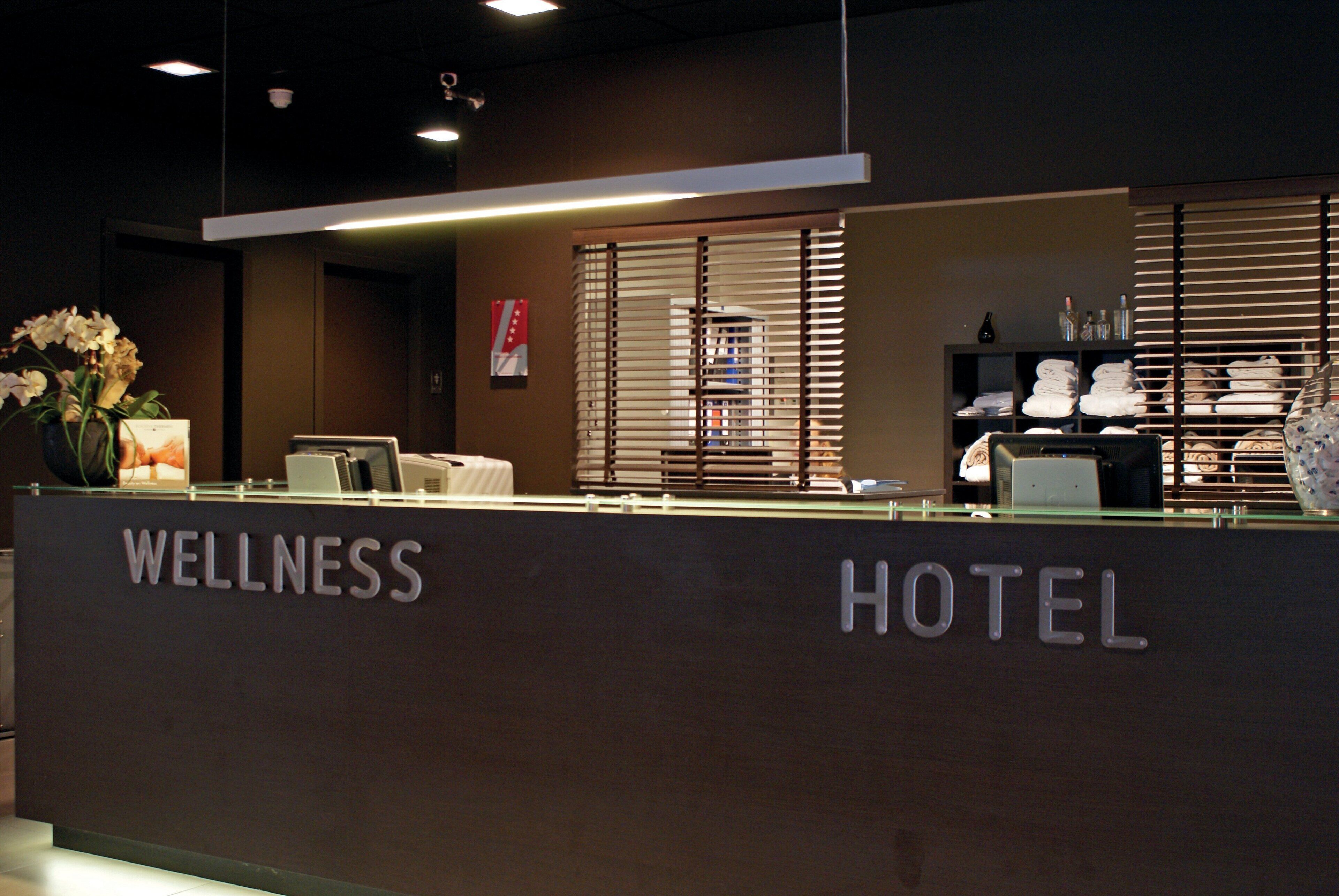 Ein Empfangsbereich mit einer Rezeption und der Beschilderung 'WELLNESS HOTEL', sowie ein Fenster mit Jalousien.