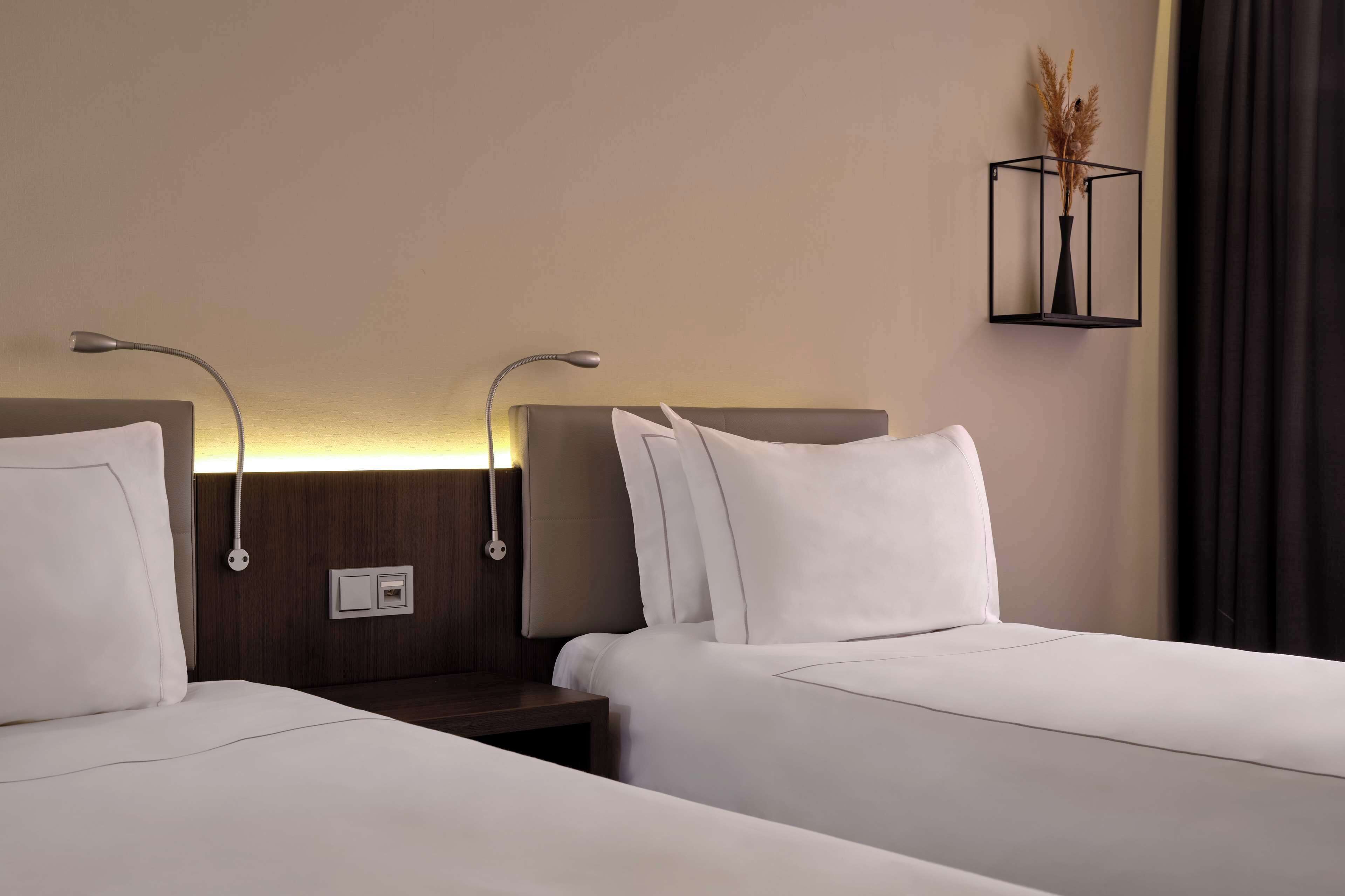Two beds with white linens, a bedside table, a wall-mounted lamp, and a decorative vase with pampas grass.