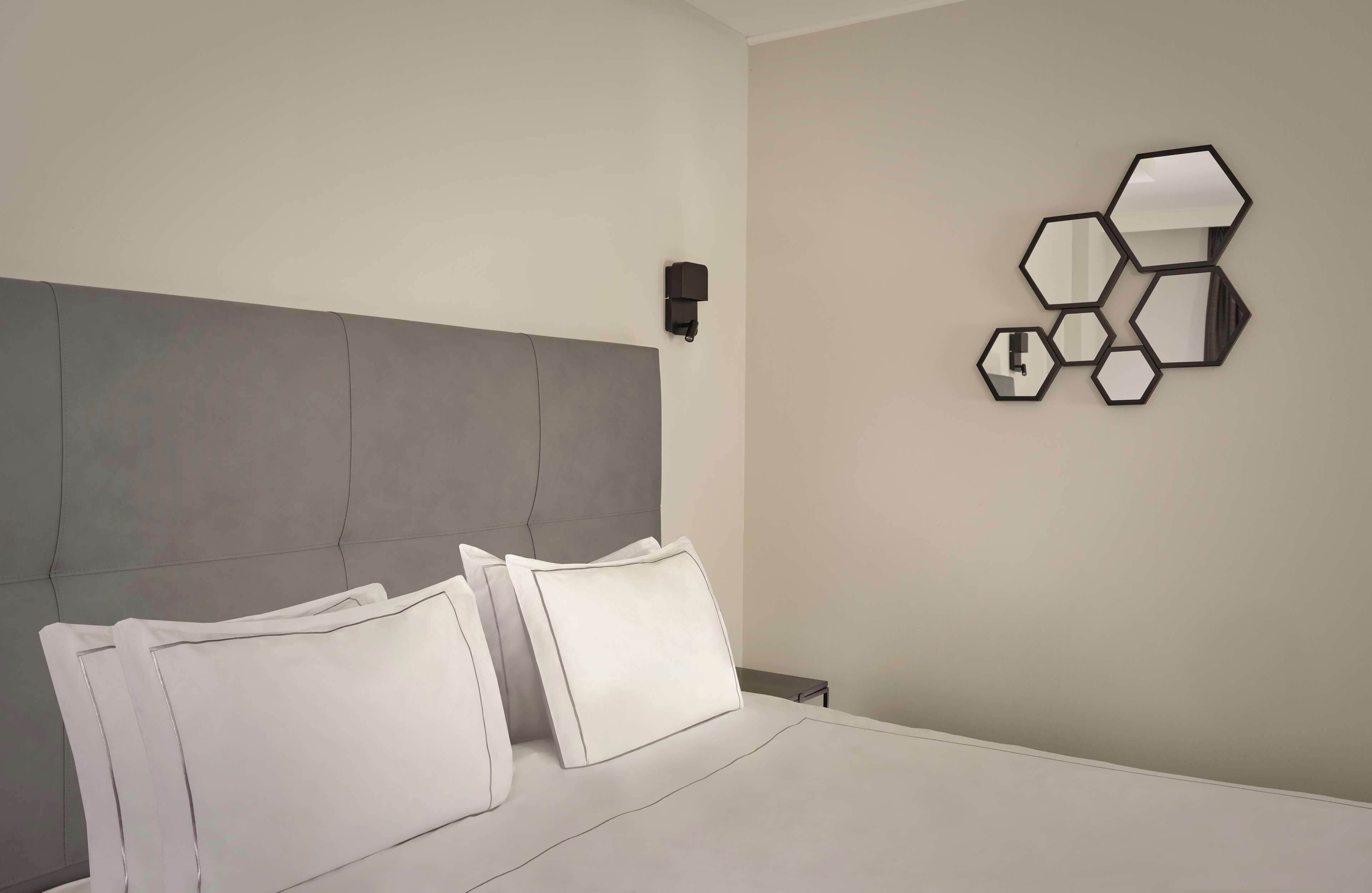 A modern hotel room with a grey upholstered headboard, a neatly made bed with white linens, and a wall-mounted hexagonal mirror arrangement.