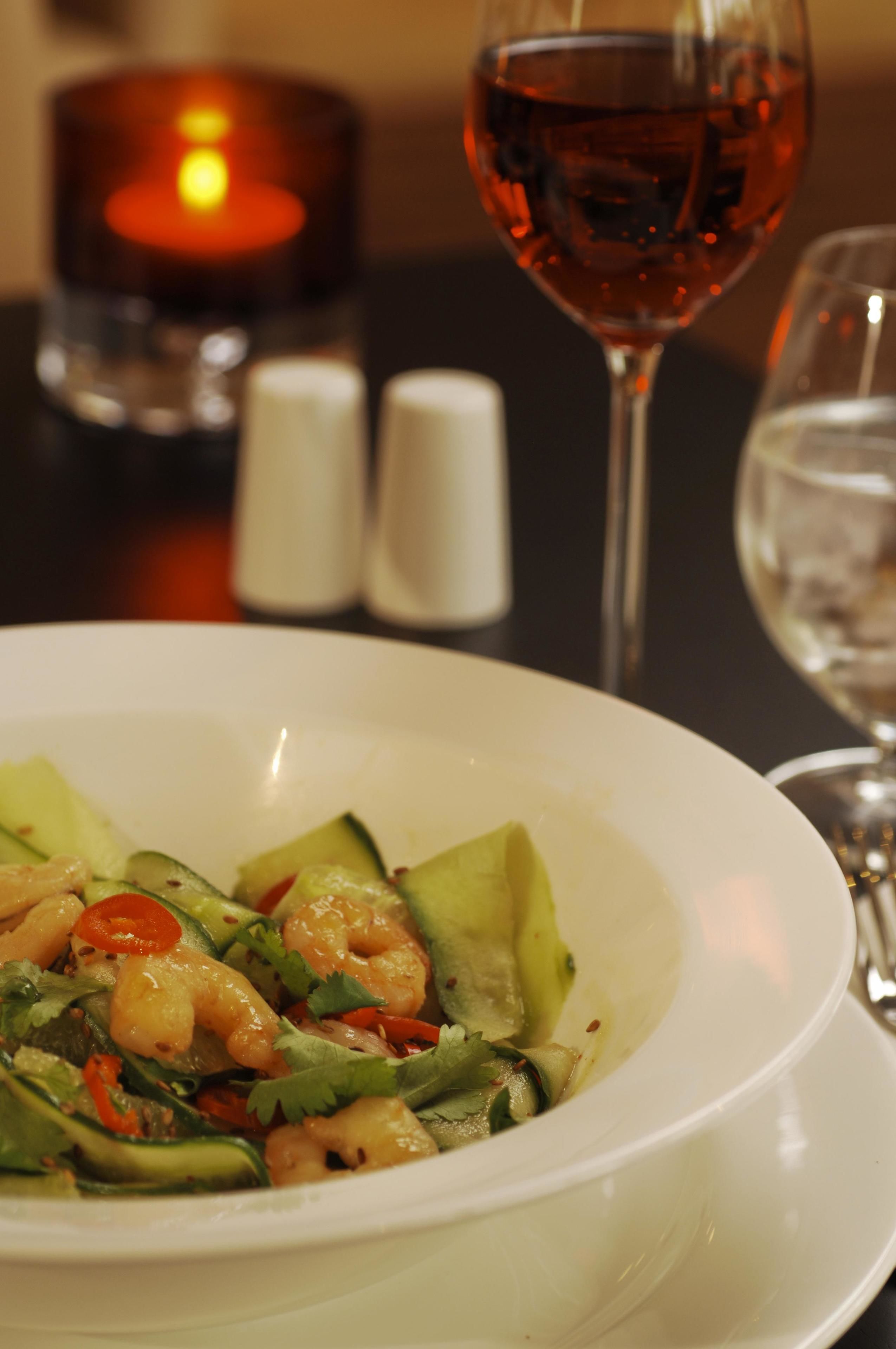 Un plato de camarones y verduras con una vela y copas de vino al fondo.