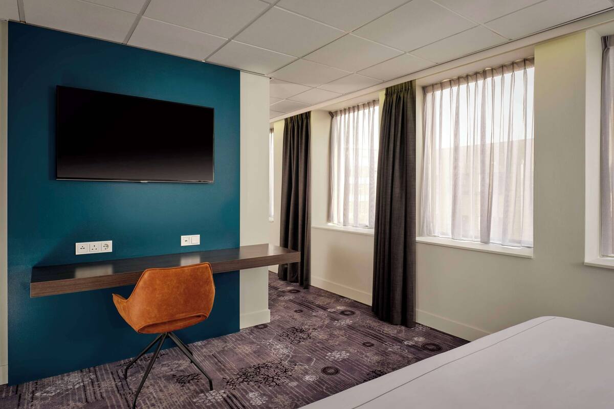 Une chambre d’hôtel dotée d’un téléviseur à écran plat fixé sur un mur de couleur turquoise, d’un bureau avec une chaise et de grandes fenêtres avec des rideaux.