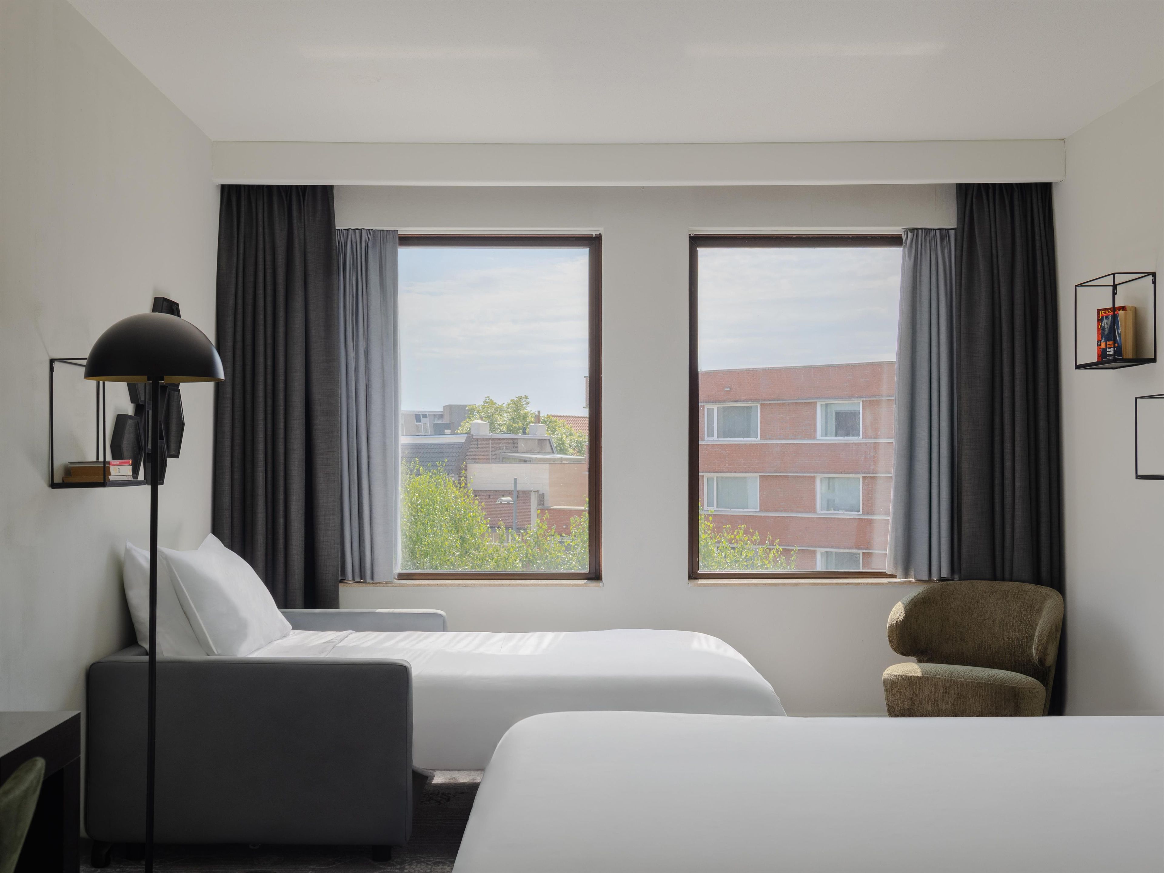 A hotel room with two beds, a desk, a chair, a lamp, and a view of buildings.