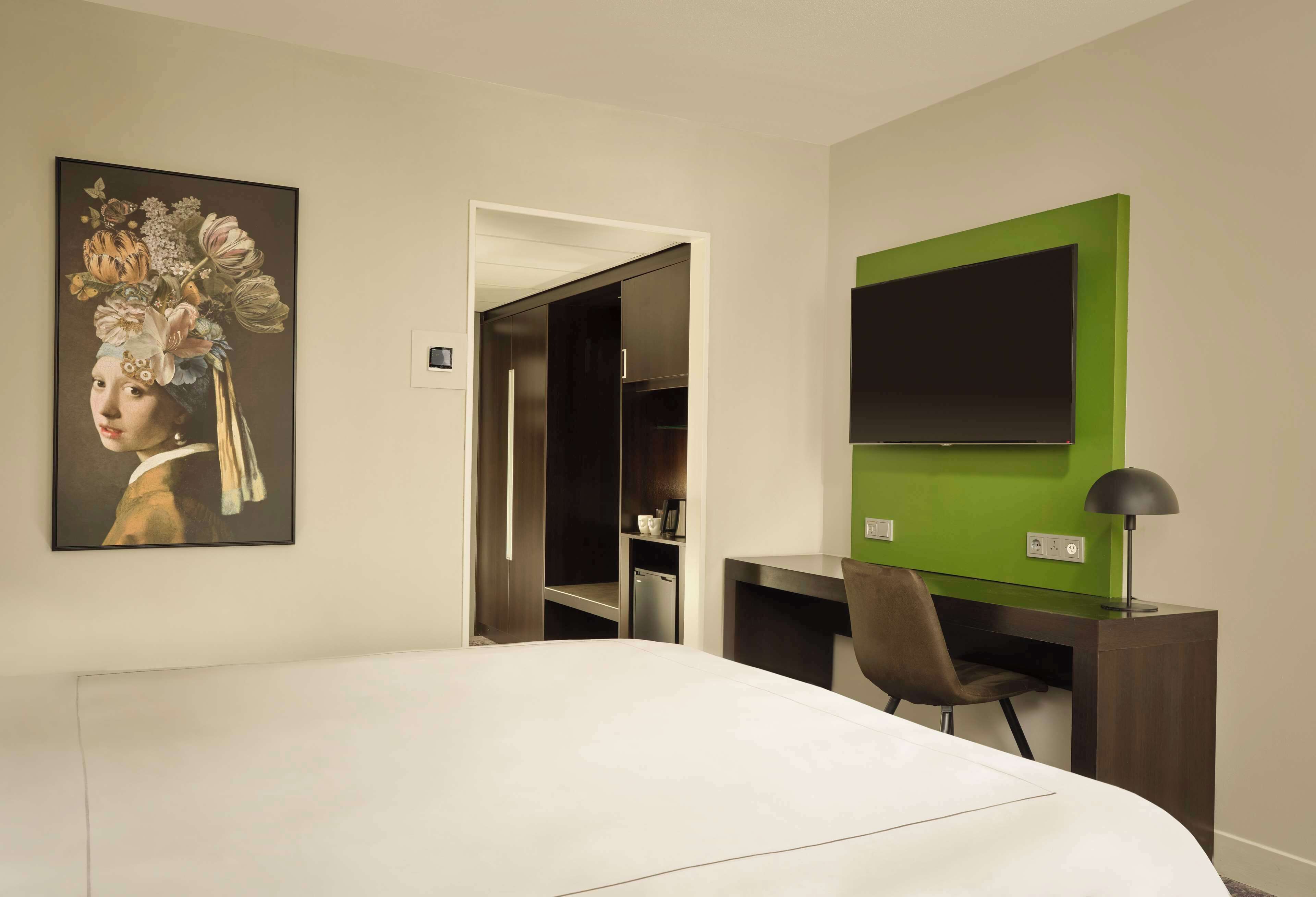 Habitación de hotel con cama, escritorio, silla, televisor y un cuadro en la pared.