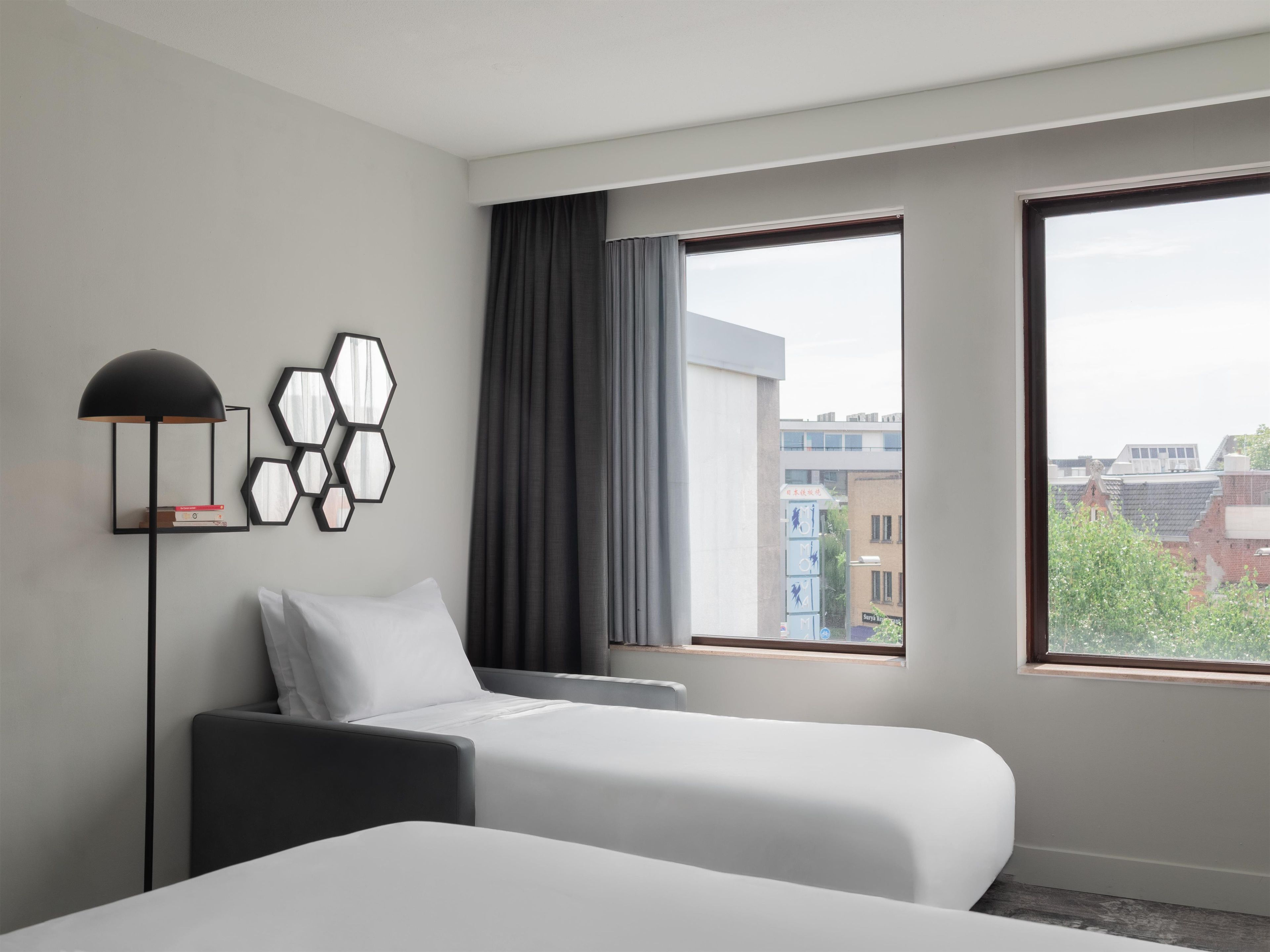 Quarto de hotel com duas camas, uma luminária de chão e vista para prédios através de janelas amplas.