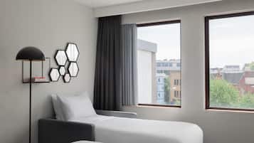 Quarto de hotel com duas camas, uma luminária de chão e vista para prédios através de janelas amplas.