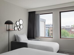 A hotel room with two beds, a floor lamp, and a view of buildings through large windows.