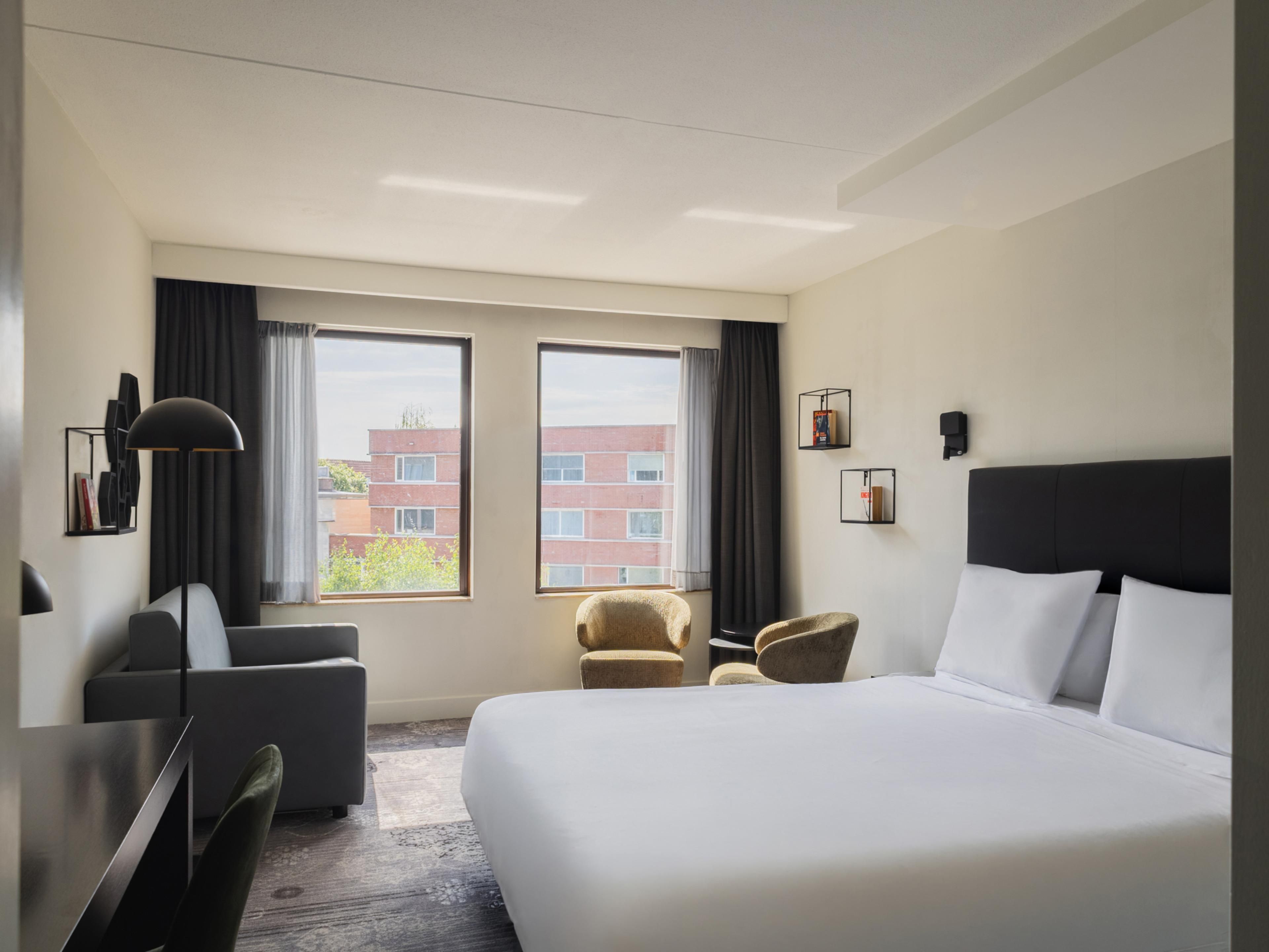 Ein Hotelzimmer mit einem großen Bett, zwei Sesseln, einem Schreibtisch und Blick auf Gebäude durch die Fenster.