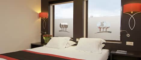 A hotel room with a large bed, two bedside tables, a nightstand, and a window with a view of buildings.