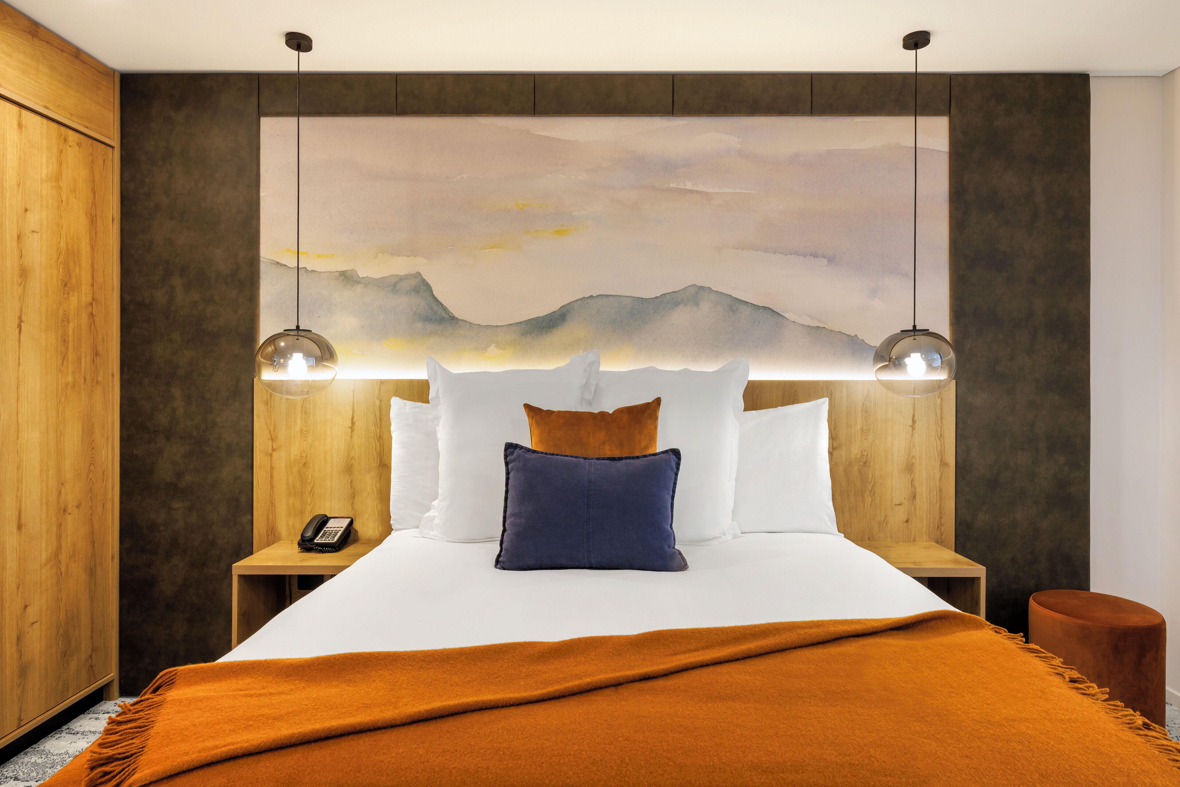 A hotel room with a large bed, a wooden headboard, a painting of mountains, and two pendant lights.