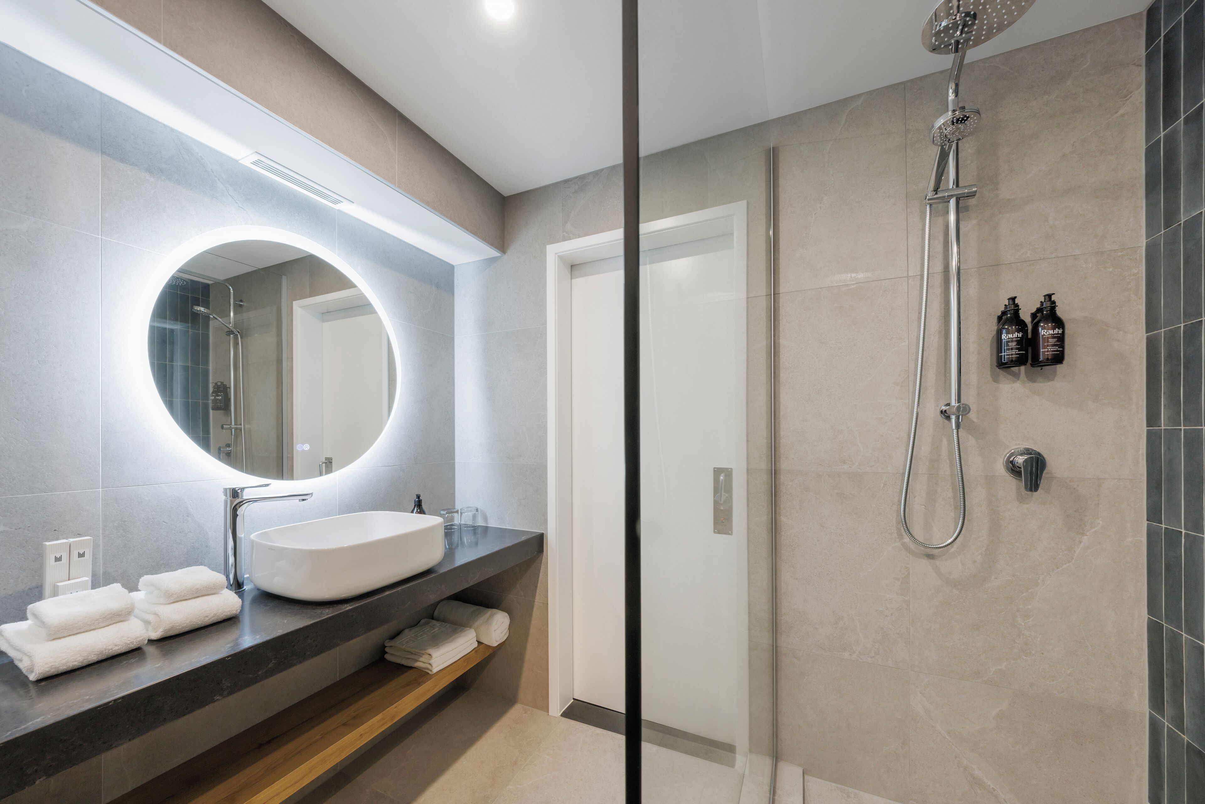 A modern bathroom with a large oval mirror, a white sink, and a glass-enclosed shower.