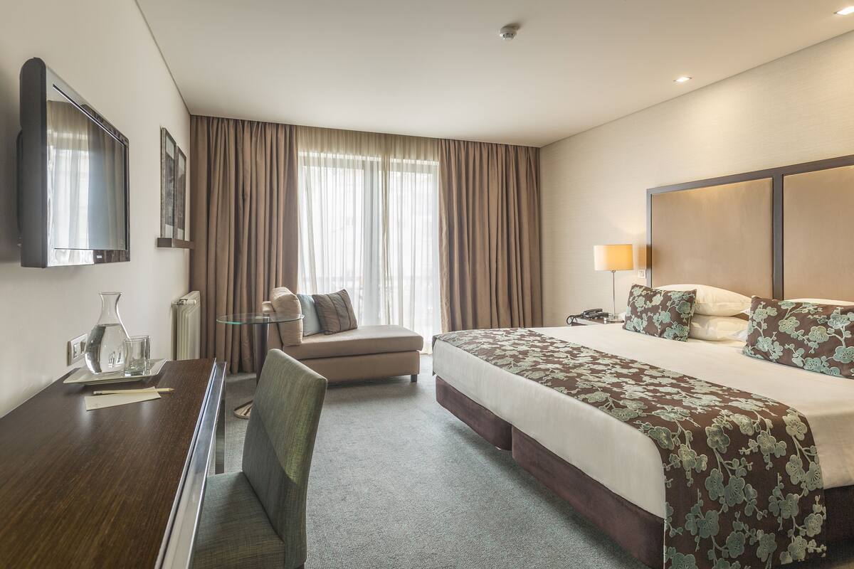 A hotel room with a bed, a desk, a chair, a small table, and a window with curtains.