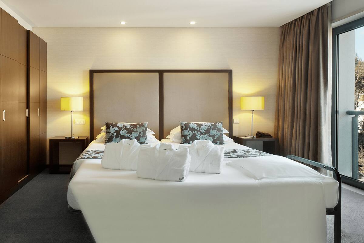 A hotel room with a large bed, two bedside lamps, a headboard, and a view of the outdoors.