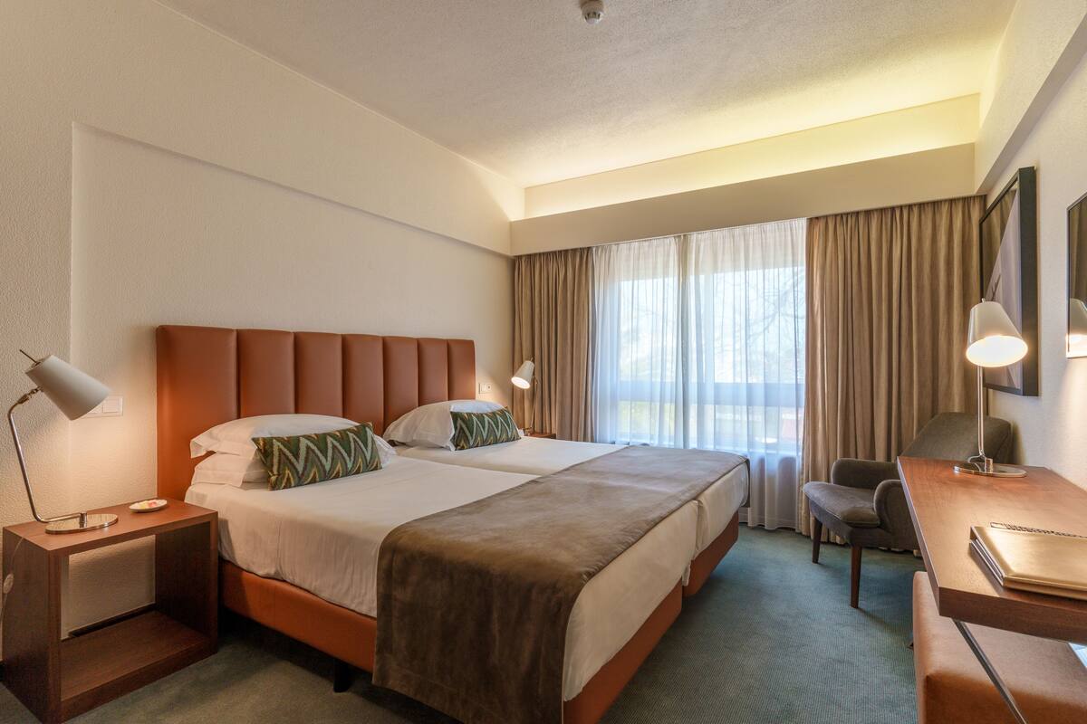 A hotel room with two beds, a desk, a chair, a lamp, and a window with curtains.