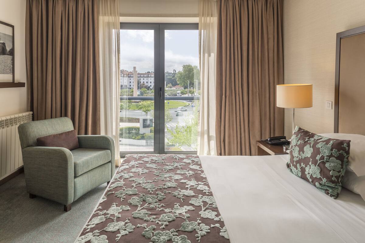 A hotel room with a bed, a chair, a lamp, and a view of the city through the window.