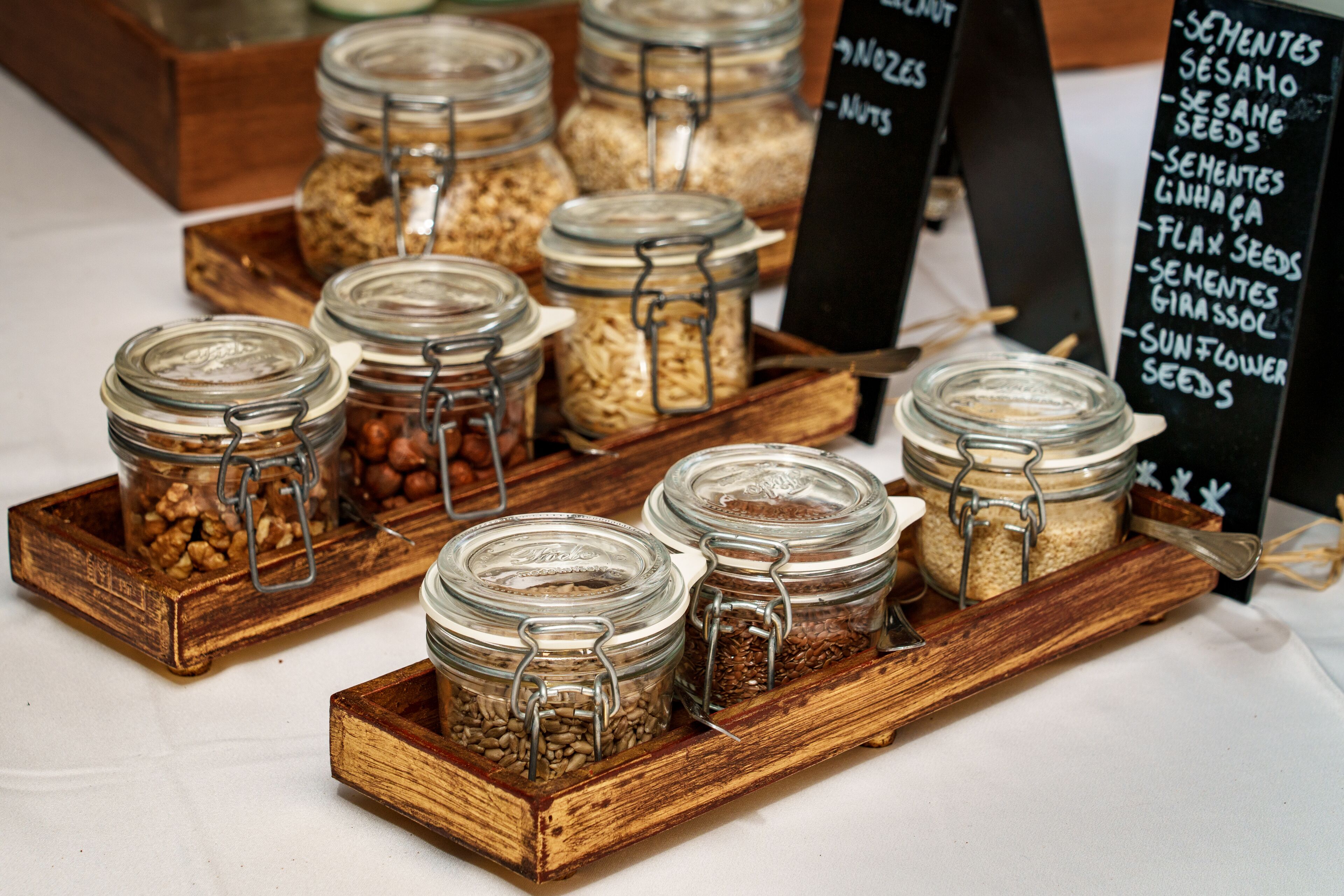 A display of various seeds and nuts in glass jars on a wooden tray.