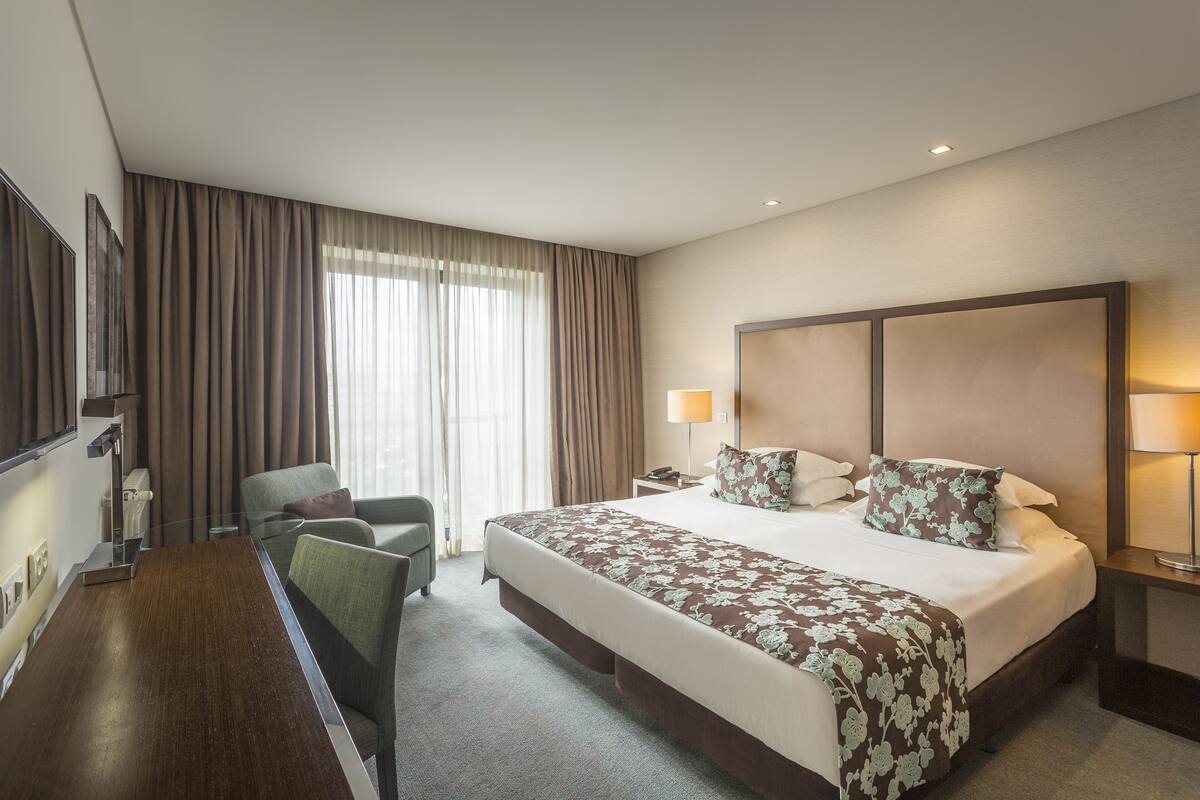 A hotel room with a large bed, a desk, a chair, a TV, and a window with curtains.