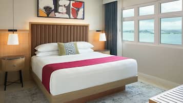 A hotel room with a large bed, two bedside tables, a headboard, a view of the sea, and a painting on the wall.