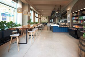A modern bar area with long wooden tables and stools, a counter with food items, and a view of the street outside.