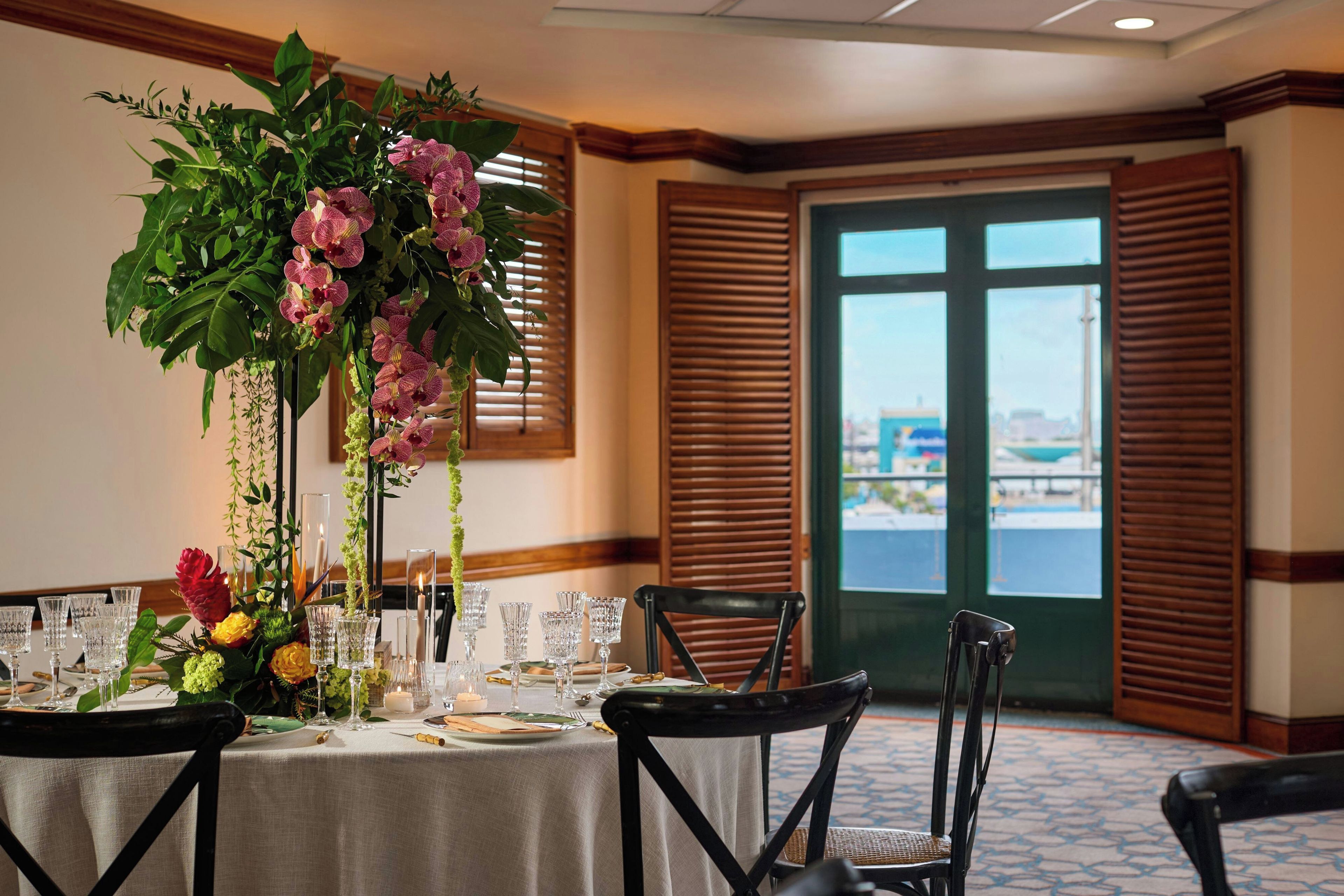 A dining area with a table set for four, featuring a floral centerpiece, clear glassware, and a view through a door with wooden shutters.