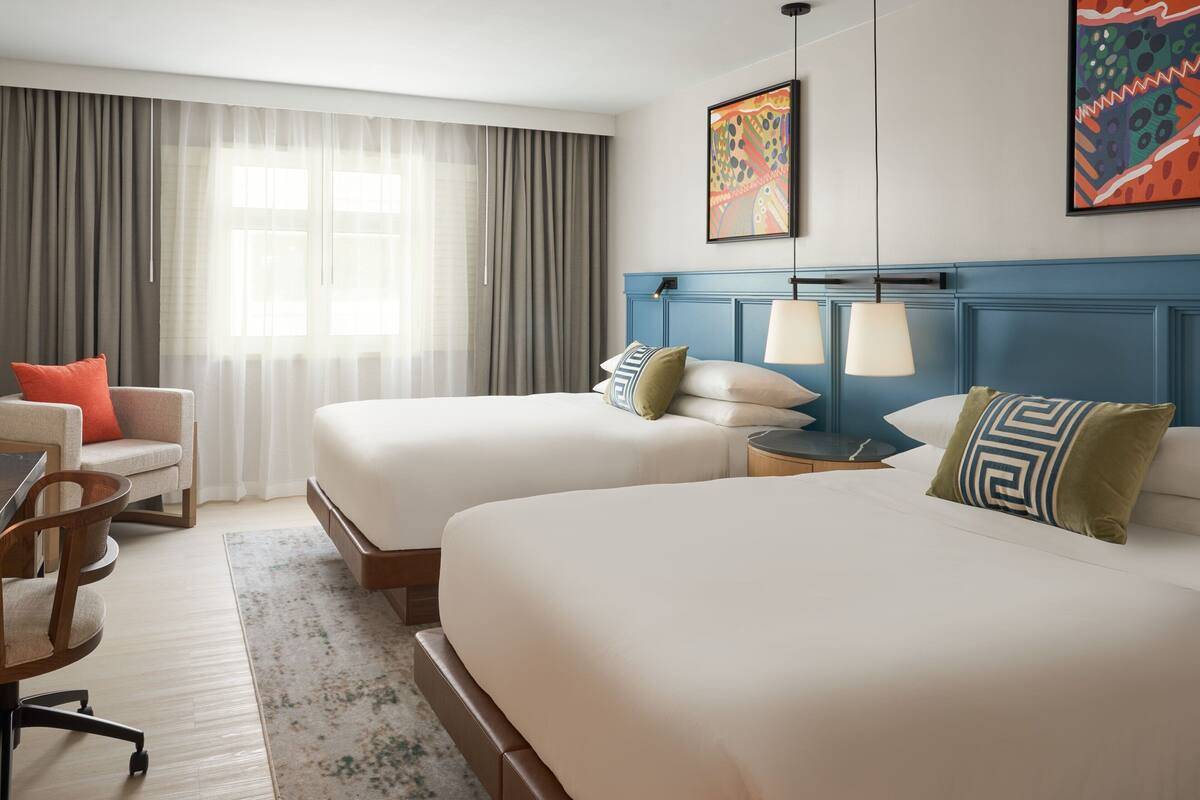 Une chambre d’hôtel avec deux lits, un bureau, une chaise et une grande fenêtre avec des rideaux.