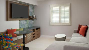 Una sala moderna con un televisor de pantalla plana montado en la pared, un sofá, un sillón tejido de colores, una mesita redonda y una ventana con persianas.