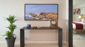 Une chambre d’hôtel moderne avec un grand tableau représentant un paysage côtier, une plante en pot, une petite table avec un haut-parleur et un lit recouvert d’une couverture rouge.