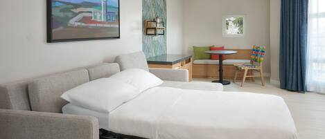 A hotel room with a bed, a sofa, a small table, and a chair. There is a painting on the wall and a window with curtains.