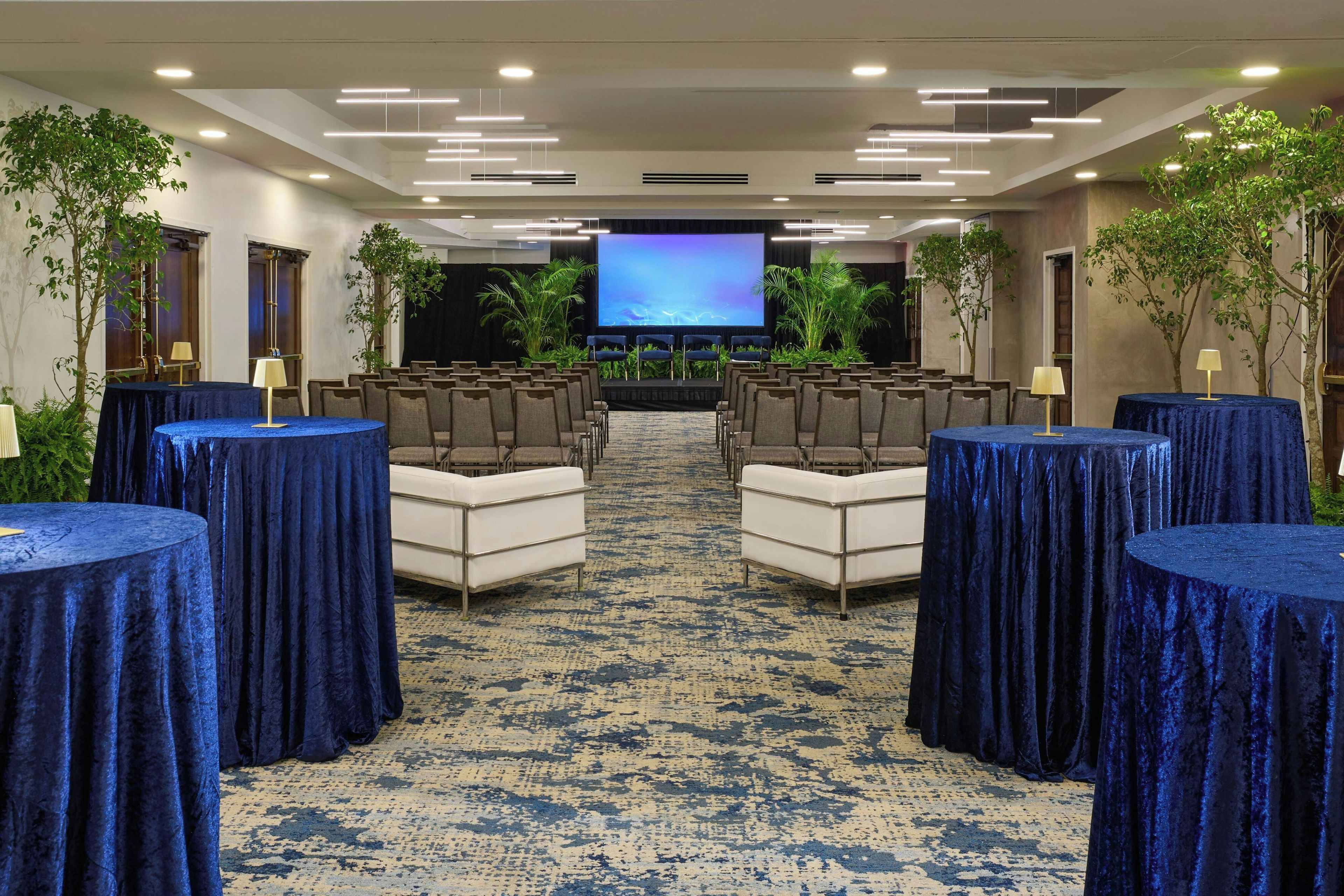 A conference room with rows of chairs, a large screen, and tables with blue tablecloths.