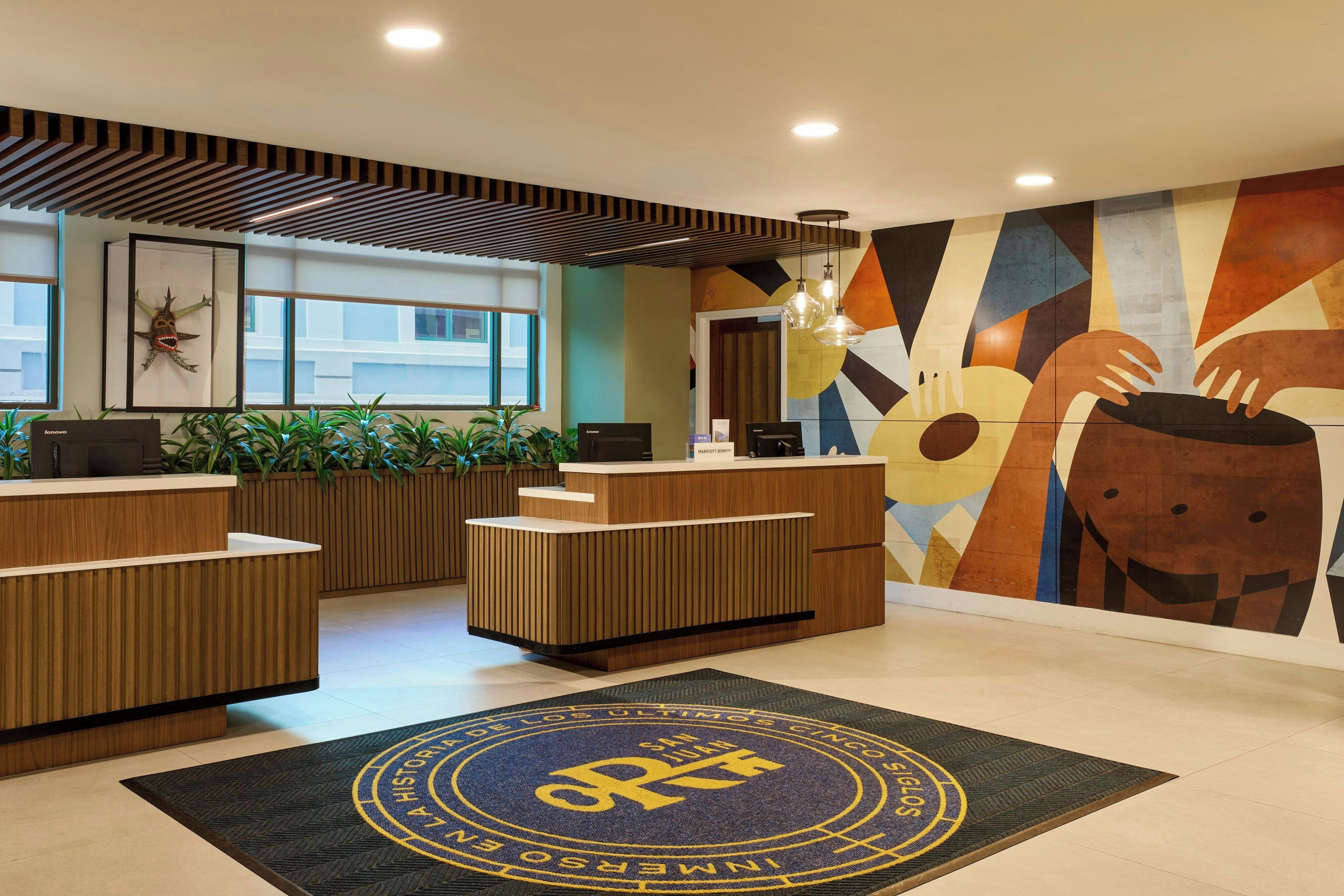 Un área de recepción de hotel con diseño moderno, que incluye un mostrador de madera, un mural grande en la pared y un felpudo con un emblema circular.