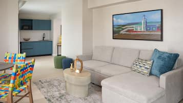 A modern living room with a grey sofa, colorful woven chairs, a small round coffee table, and a painting of a lighthouse on the wall.