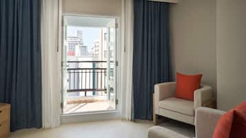 A modern living room with a sofa and a chair, a sliding glass door leading to a balcony, and a view of the cityscape.