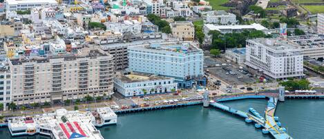 Un paisaje urbano costero con una fortaleza histórica destacada, un hotel moderno de fachada azul y un muelle frente al agua.