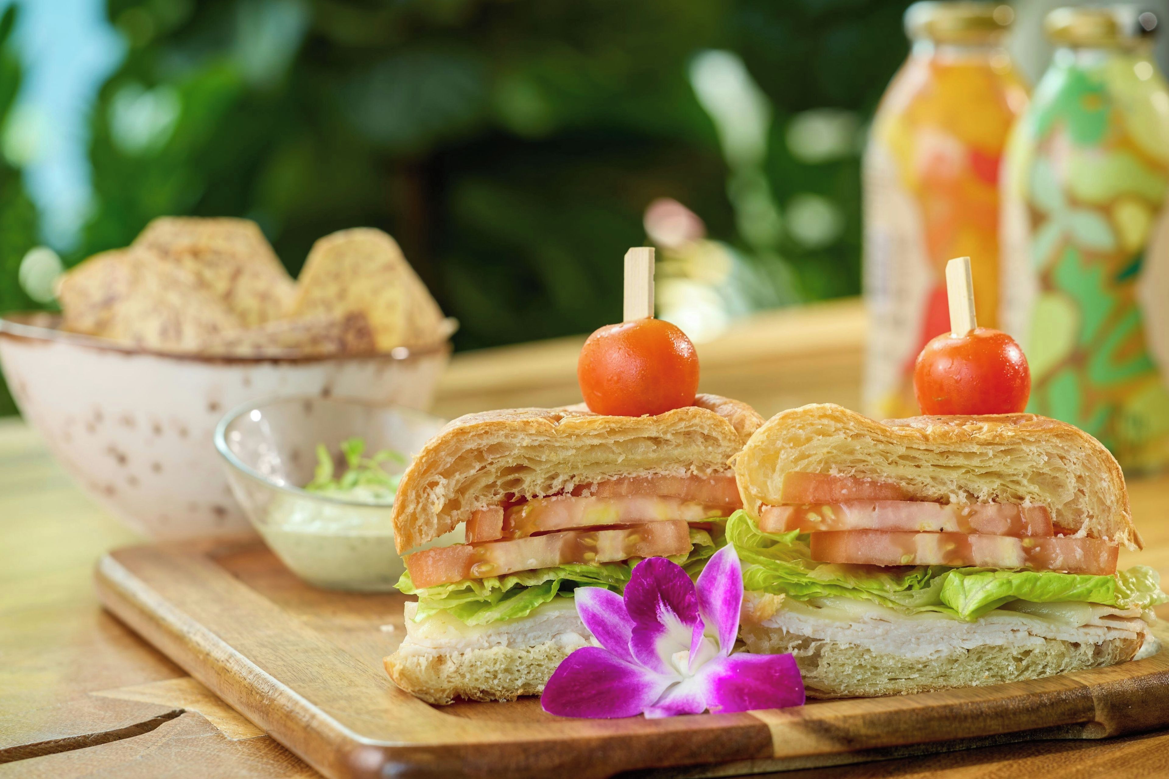 Un sandwich avec une tomate enfilée sur une brochette, un bol de frites et une bouteille de sauce sur une table en bois.