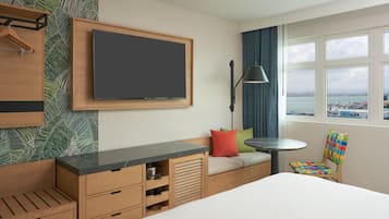 A hotel room with a bed, a TV mounted on the wall, a small table, a chair, and a window with a view of the sea.