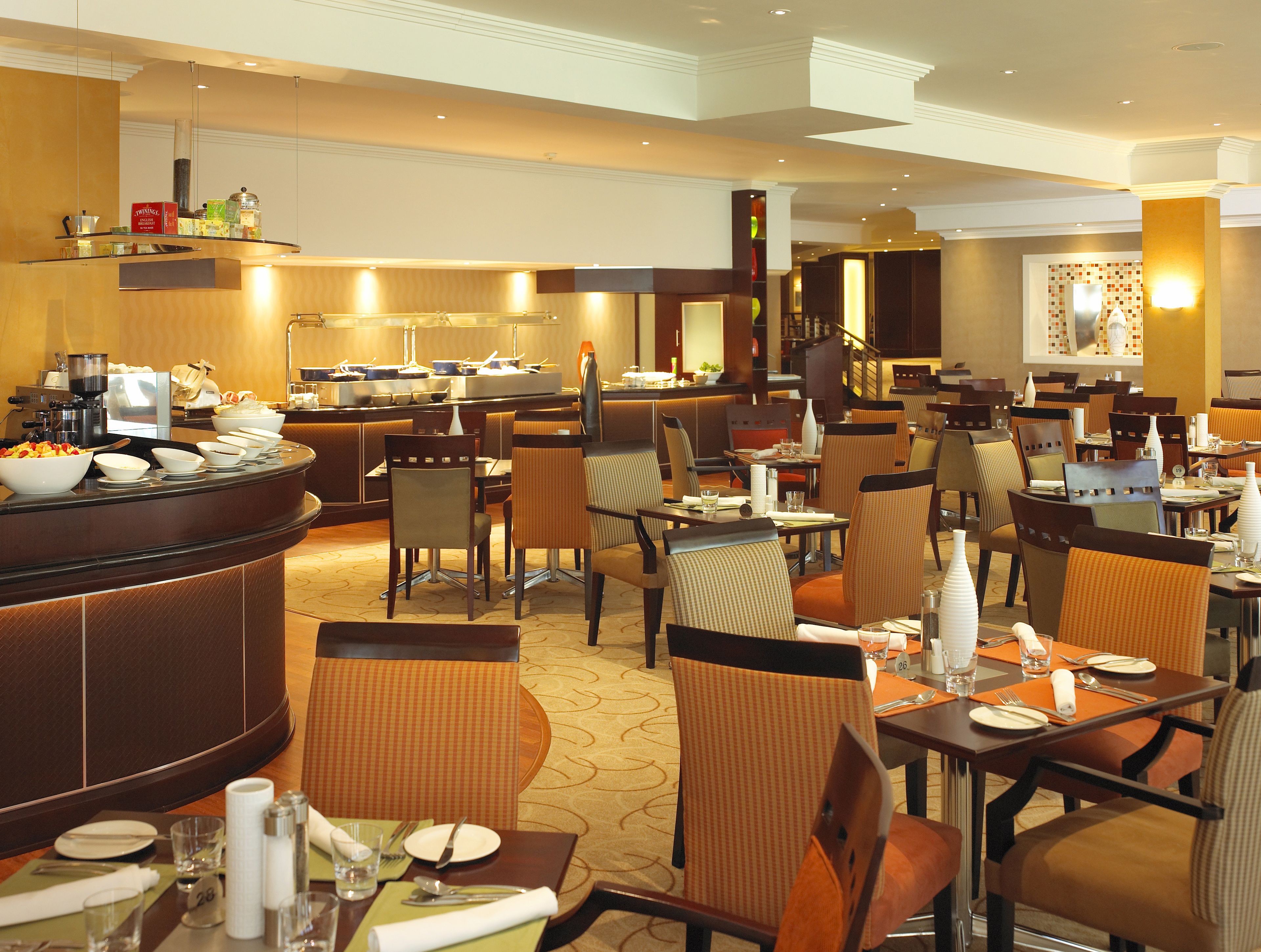 A hotel restaurant with multiple dining tables and chairs, a buffet area with various food items, and a kitchen in the background.