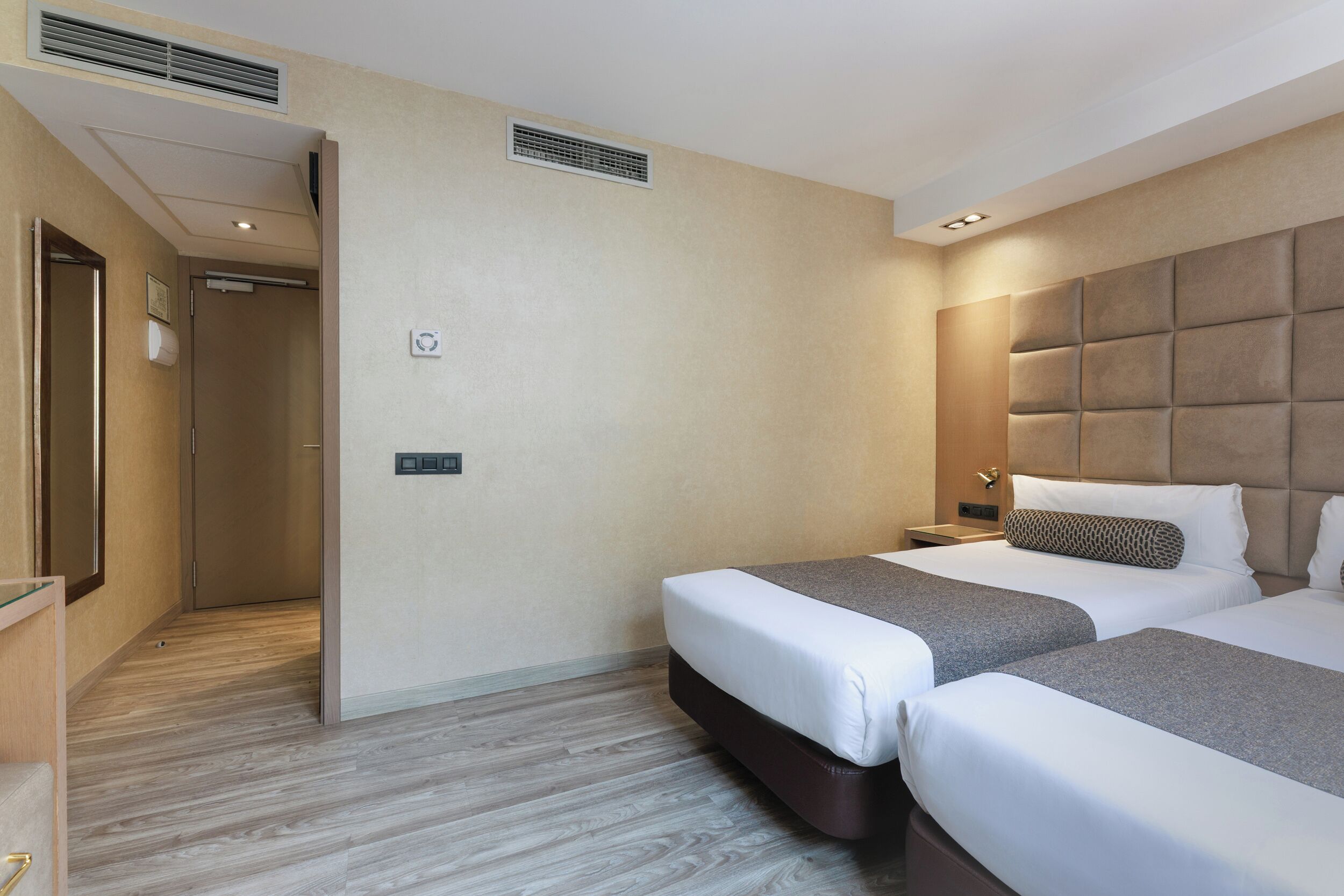 Habitación de hotel con dos camas, suelo de madera, pared color beige y diseño moderno.