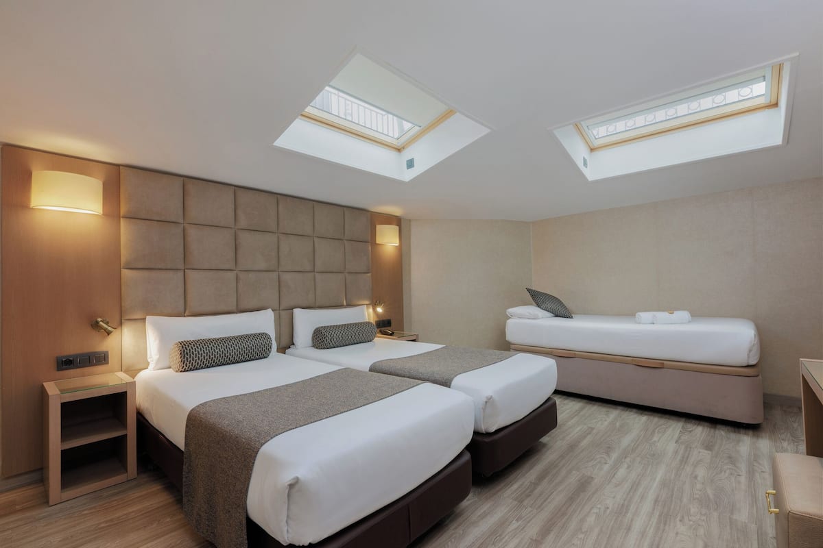 Ein modernes Hotelzimmer mit zwei Betten, einer Schlafcouch, einem Oberlicht und einem kleinen Schreibtisch.