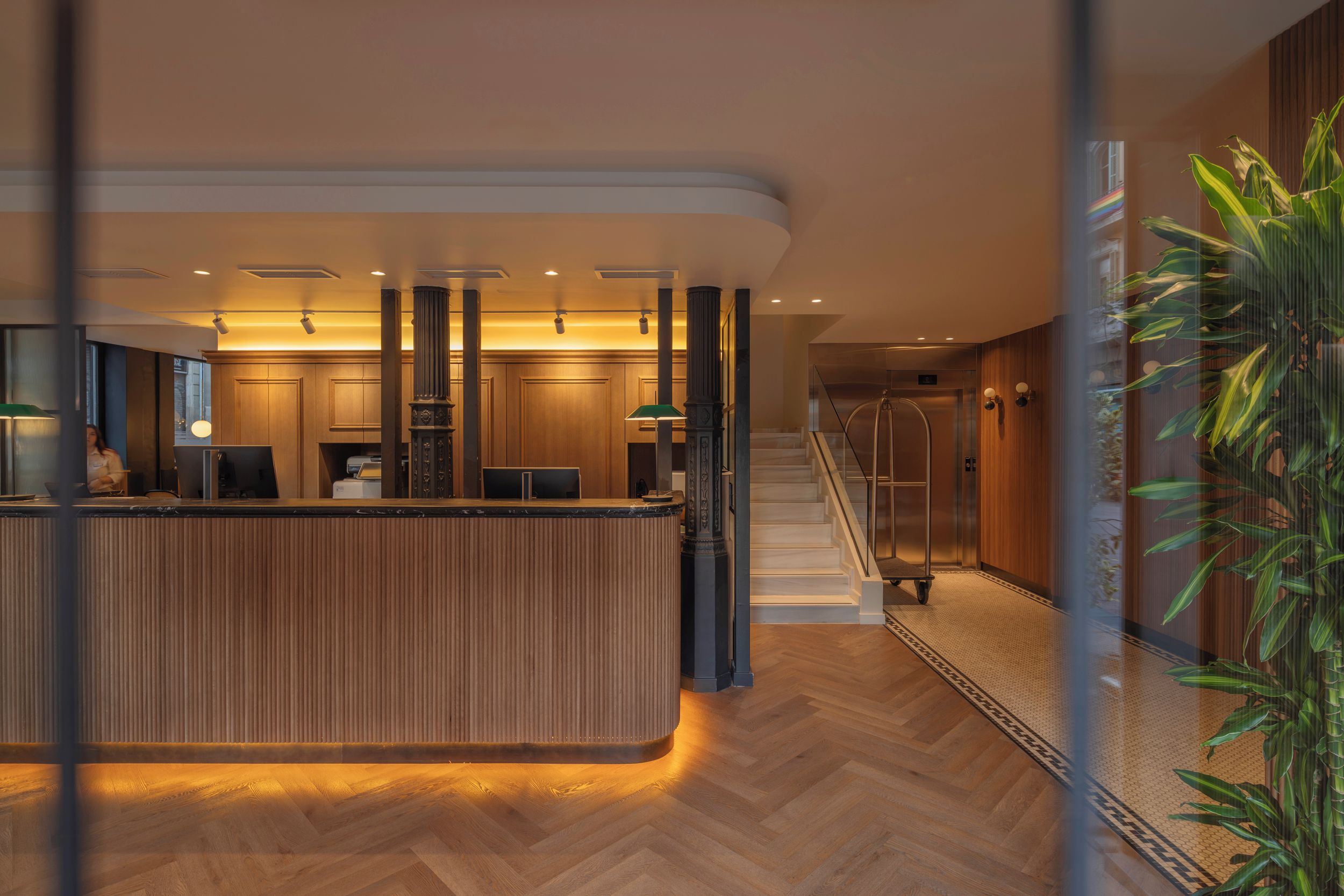 Zona de recepción de hotel con un mostrador de madera, una escalera y un panel de vidrio.