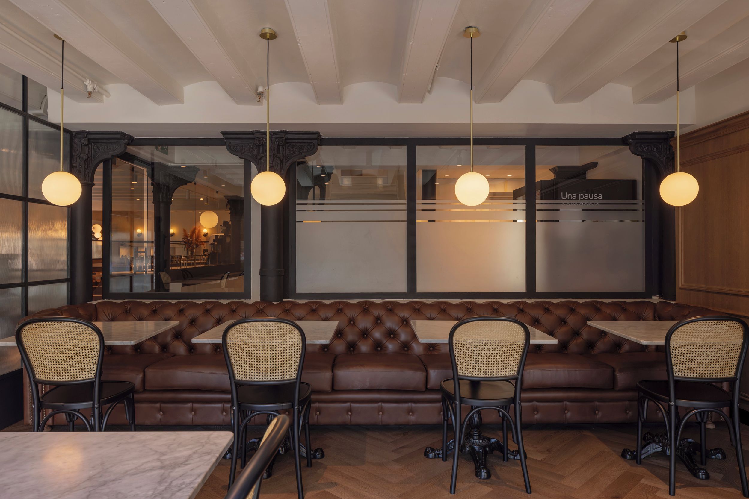 Un interior de restaurante con un largo banco de cuero marrón, cuatro mesas con sillas e iluminación colgante.