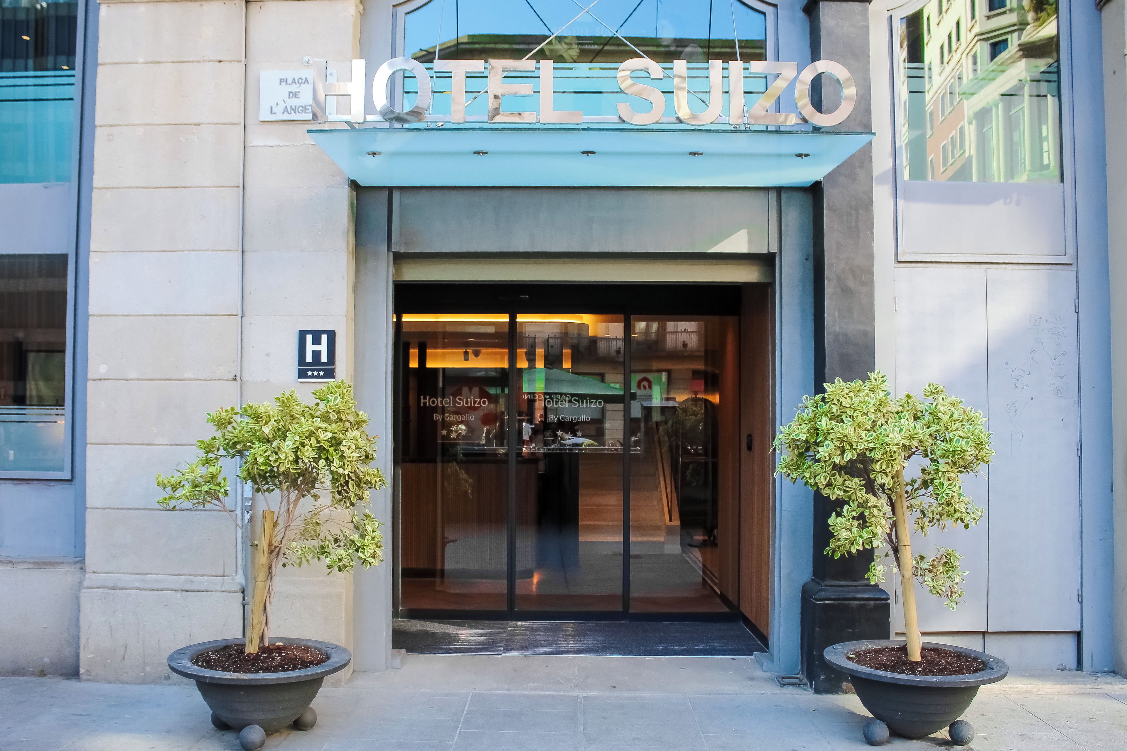 Der Eingang zum Hotel Sulzo mit einem Schild über der Tür, einem Empfangstresen im Inneren und zwei Kübelpflanzen im Vorbereich.
